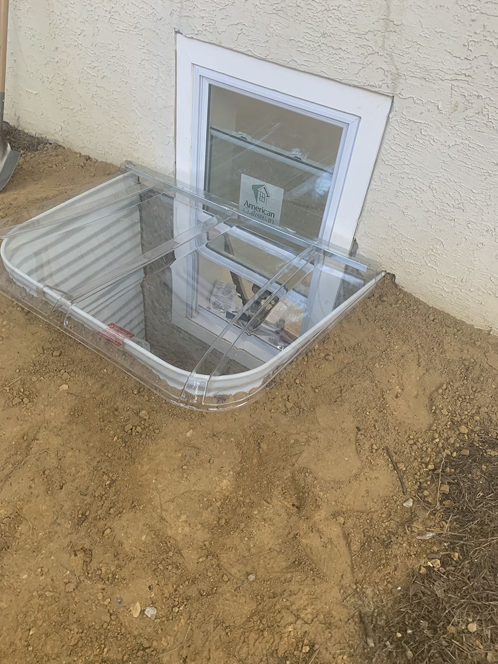 A window well is sitting in the dirt next to a house.