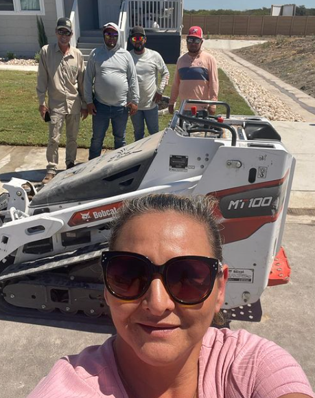 woman taking selfie with group near Bobcat MT 100 and house; men in background