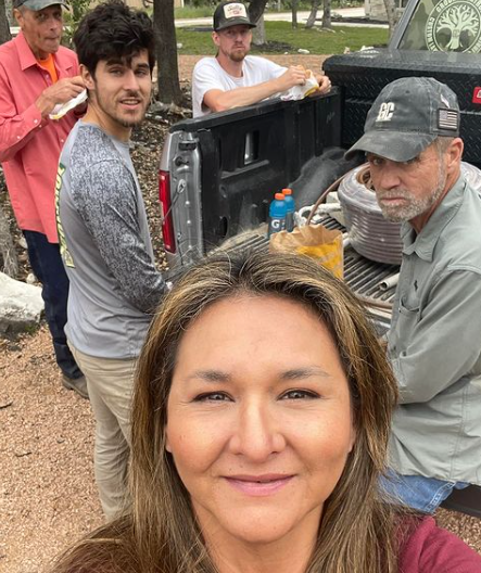woman takes selfie with group by a truck, preparing food outdoors
