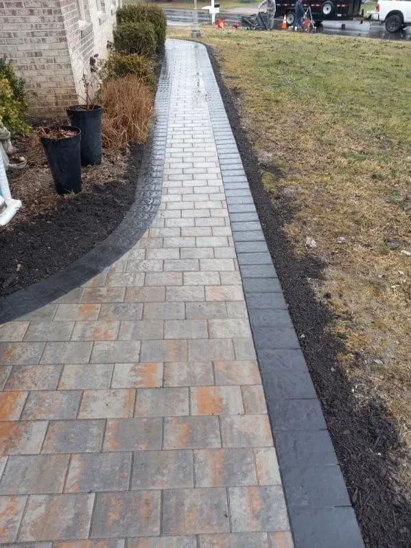 Brick walkway with dark borders next to a grassy lawn and landscaped flowerbeds.