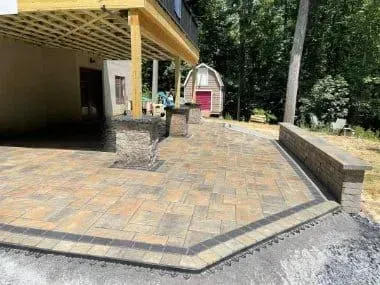 Stone patio with retaining wall, under a wooden deck, near a shed and trees.
