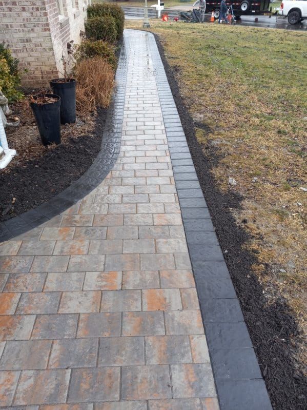 Brick walkway with dark border next to grass and mulch; pots with plants on left.