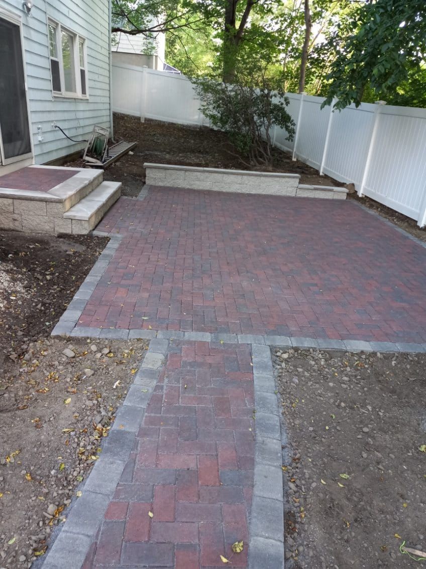 Brick patio and walkway with a raised garden bed, next to a house with white fence.