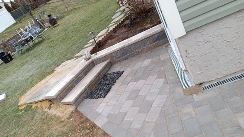 Stone patio with steps, wall, and drainage next to a house with green siding. Grass yard in the background.