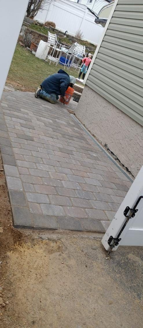 A person kneels, cutting pavers with an orange saw, on a stone path. Gray siding and white fence in the background.