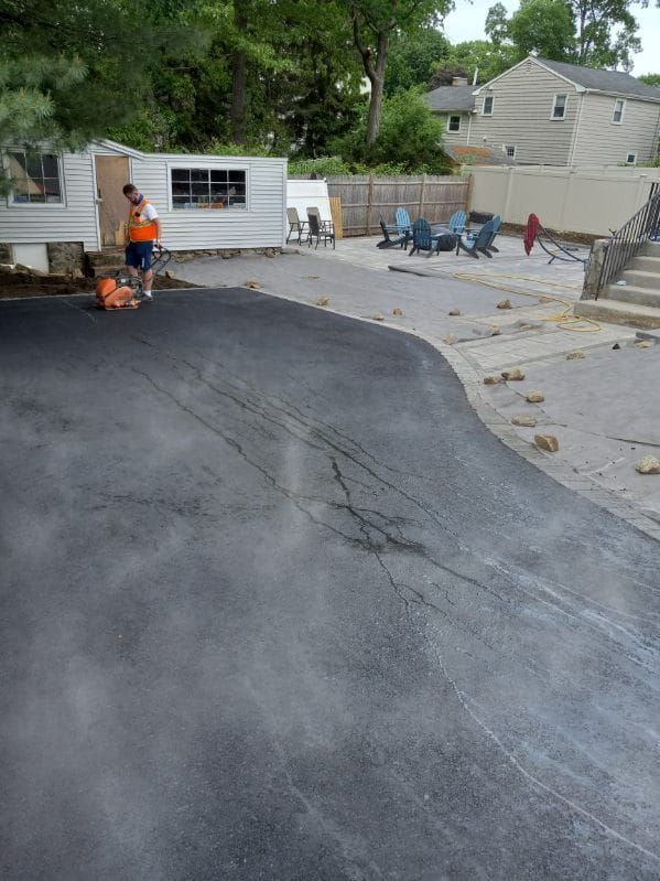 Man paving driveway; gray asphalt, cracks visible. Houses and yard in background.