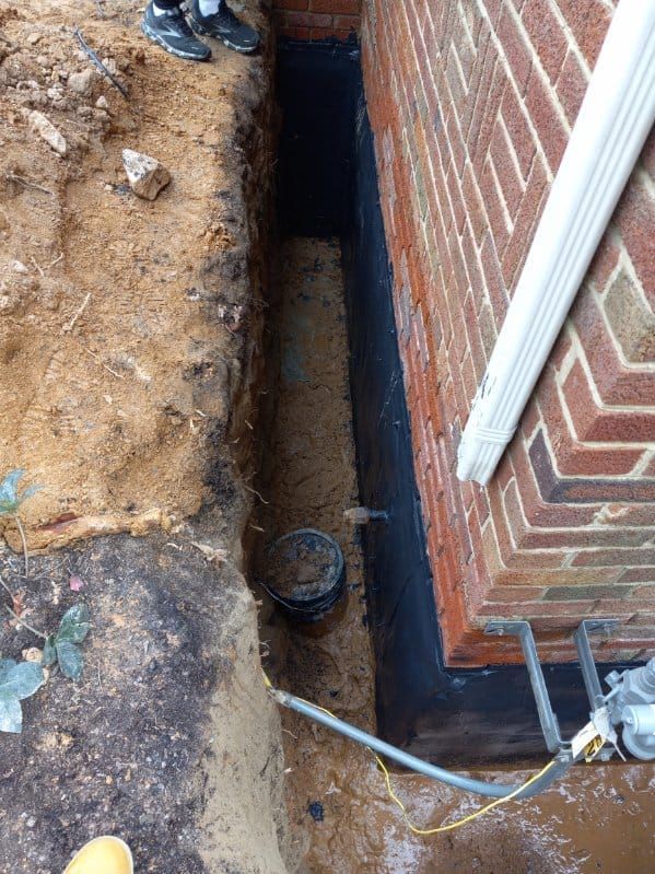 Trench dug next to a brick wall. Black sealant on the wall. Person's legs visible at top.