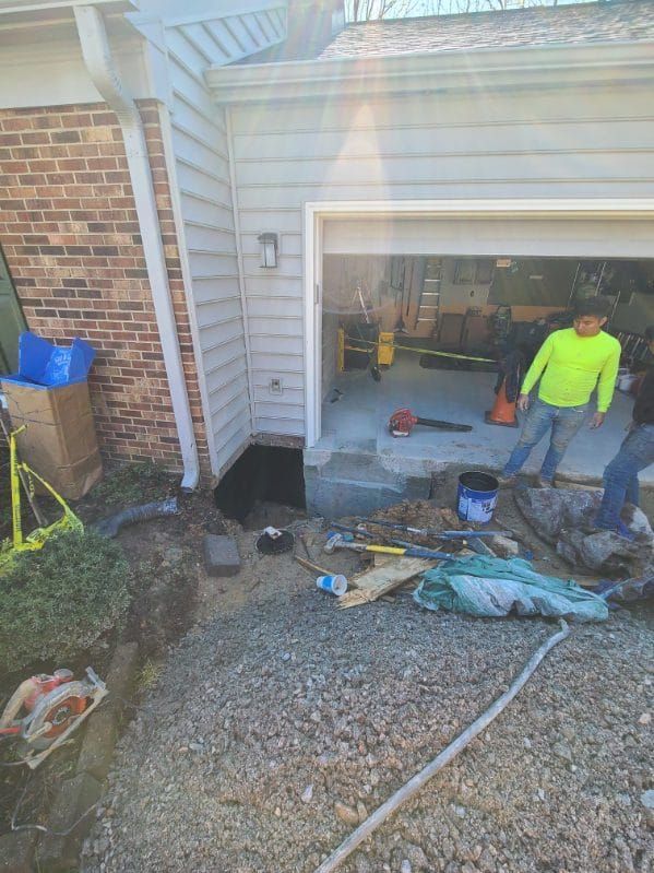 Construction site: Crew working on garage entrance, exposing foundation. Gravel, tools, and soil surround a black opening.