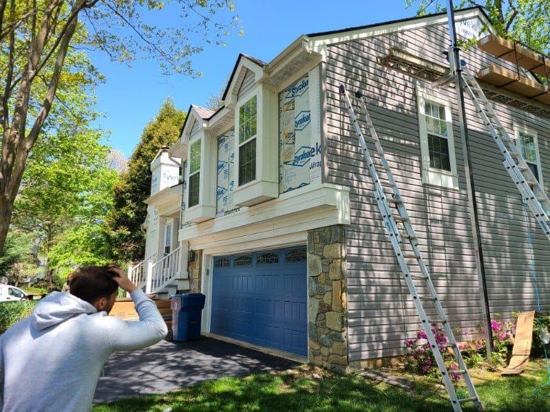 Exterior of a house under construction; man looking at the facade. Gray siding, blue garage door, ladders, and scaffolding.