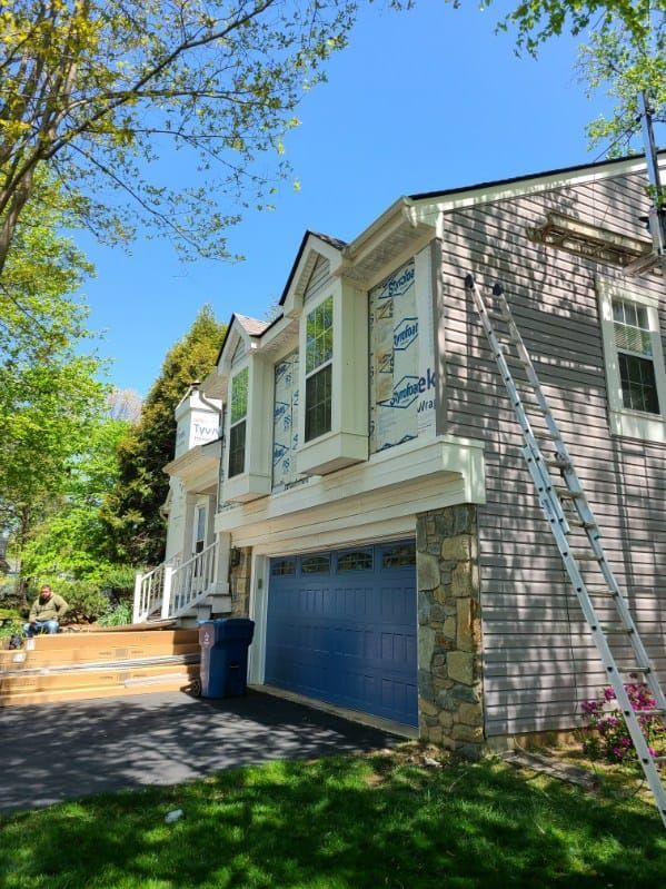Two-story house undergoing renovations. Gray siding, blue garage door, blue wrap visible, ladder. Sunny day.