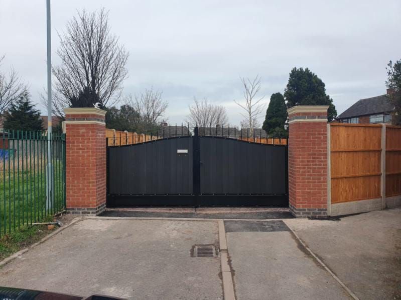 Black gate between brick pillars, opening onto a driveway, with a wooden fence to the right.