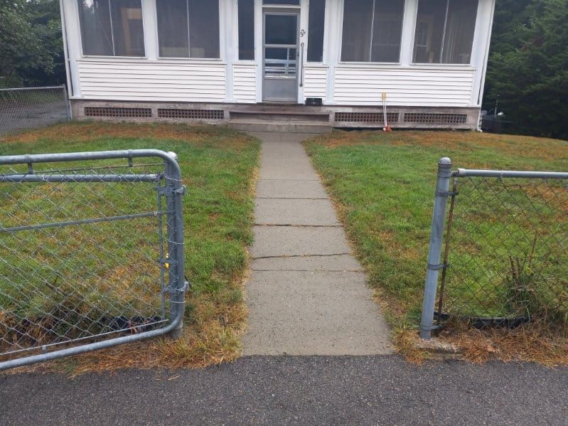A concrete walkway leads to a white house with a screened porch. Two open metal gates flank the path.
