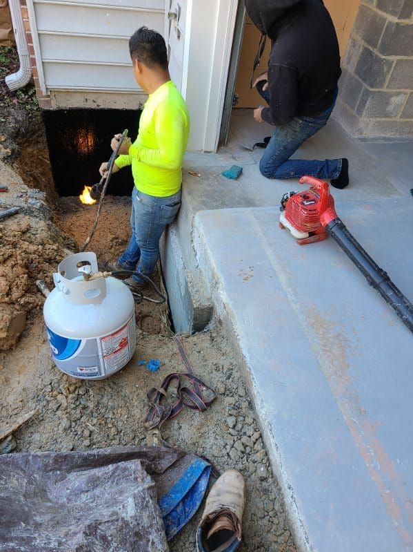 Two men work on a building's foundation. One uses a torch, the other kneels with a leaf blower.