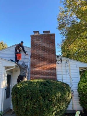 Two people on a ladder, one on a roof, painting the side of a house, near a brick chimney.