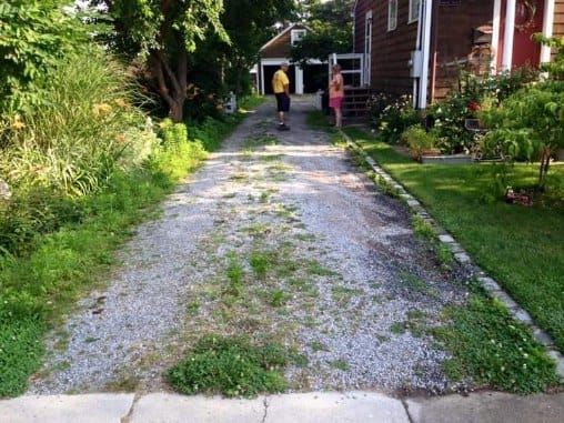 Driveway overgrown with weeds, two people stand near the garage of a house.