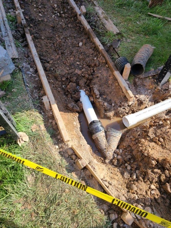 Trench with exposed white PVC pipe, connecting to a perforated drain pipe. Yellow caution tape on the ground.