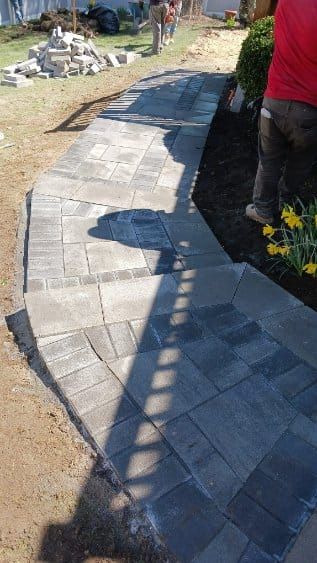 Workers laying brick pavers for a walkway in a yard, with soil and landscaping visible.