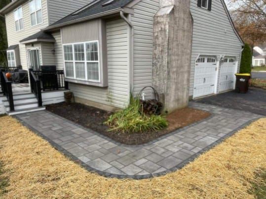 Grey paved walkway curves around a house with a chimney and garage.