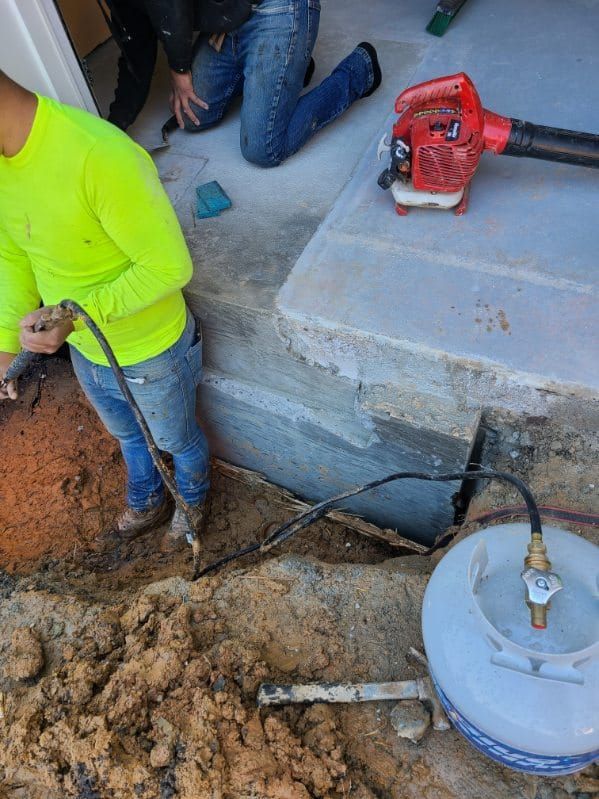 Workers using tools near a concrete structure. A gas tank and leaf blower are visible; one person in neon green.