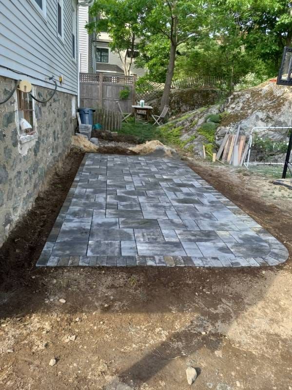 Stone paver patio under construction next to a house and hillside. Earthy tones and gray pavers.