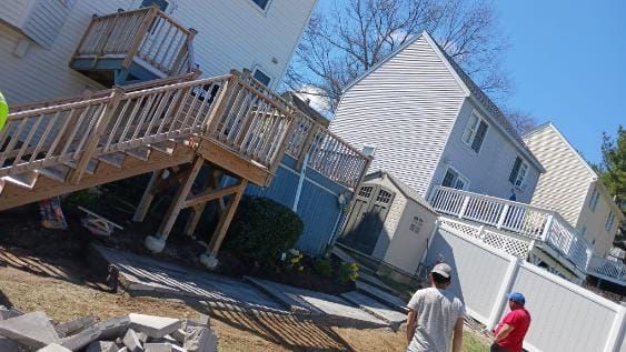 A house with wooden decks, stairs, and two people standing in a yard on a sunny day.