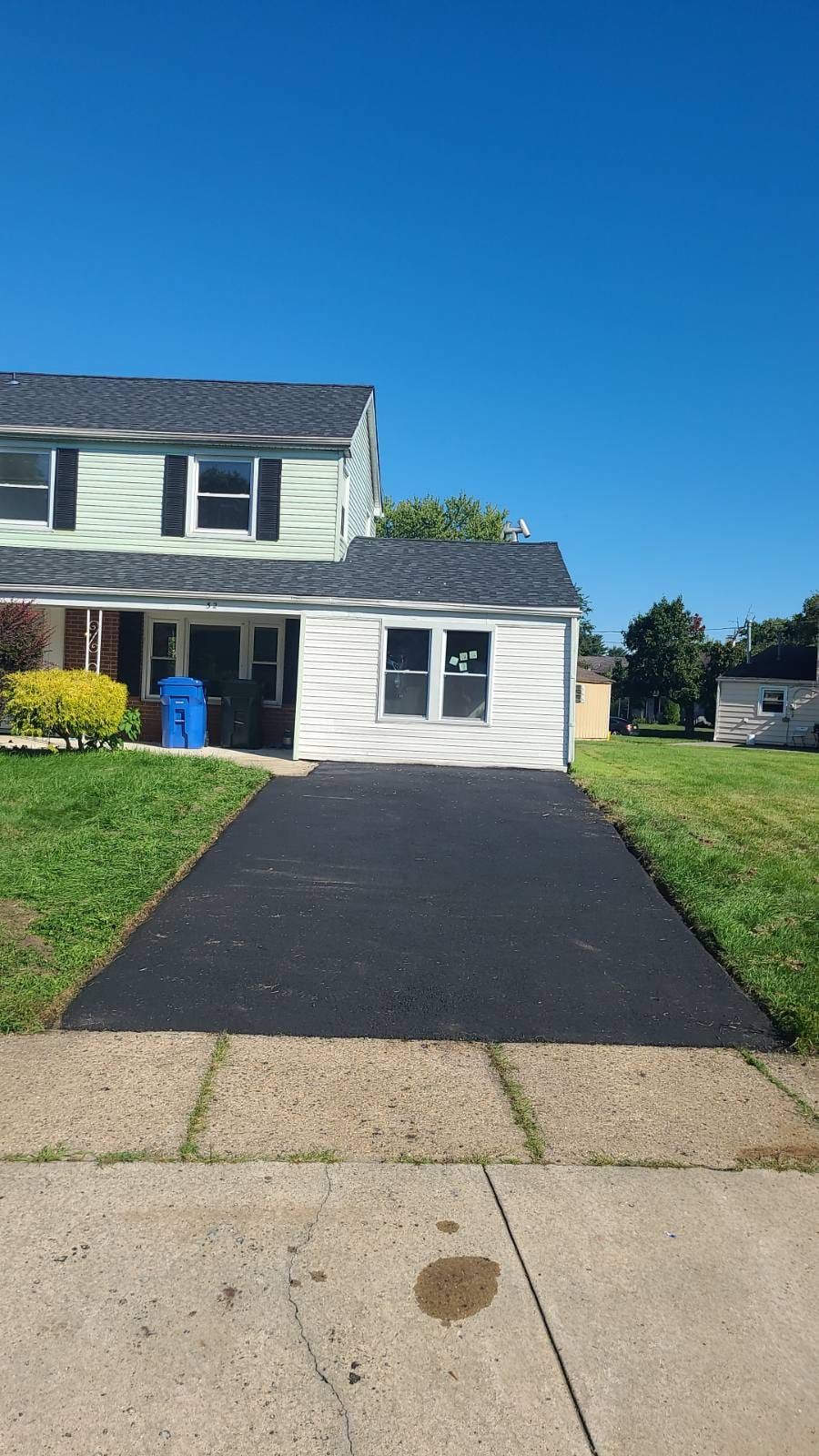 Two-story light green house with black driveway under a bright blue sky.