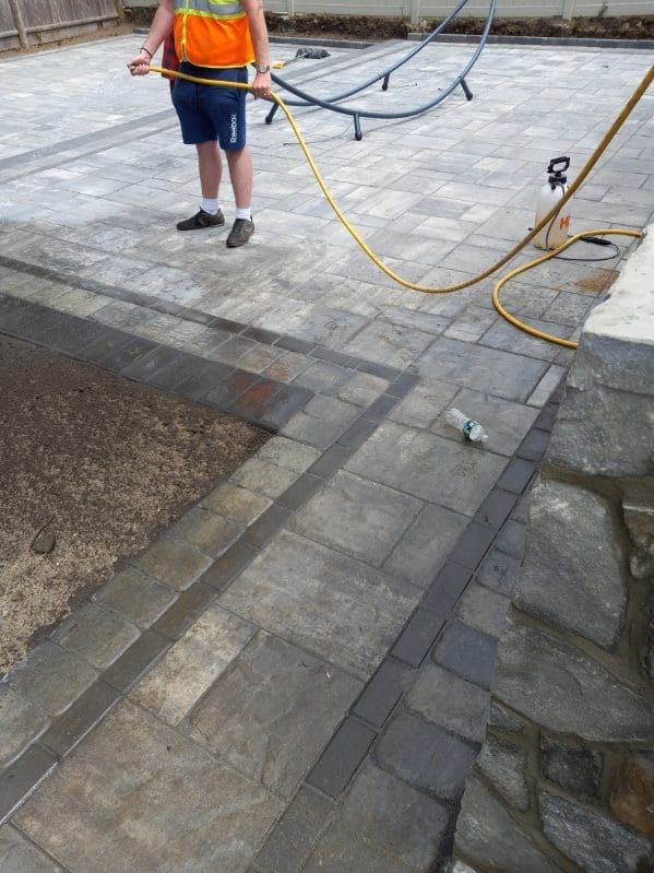 Person in orange vest sprays a stone patio with a hose; patio has steps and borders.