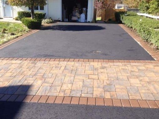 A black asphalt driveway with a brick paver apron, bordered by hedges and a garage.