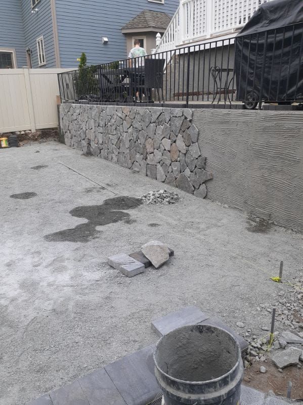 Stone retaining wall under construction in a yard with gravel and concrete.