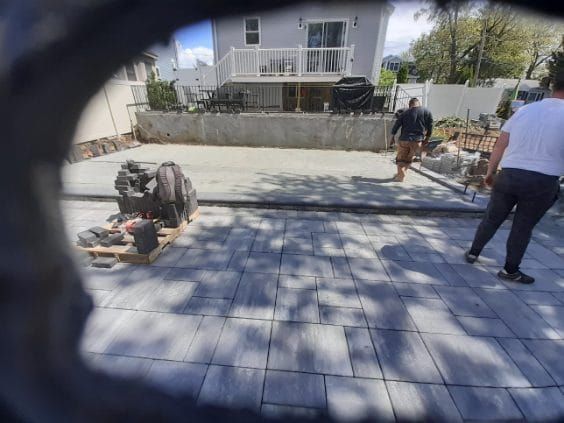 Workers paving a backyard patio with gray rectangular pavers, near a house with a deck.