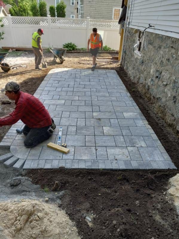 Workers laying grey paving stones for a patio; one kneels, two others walk.