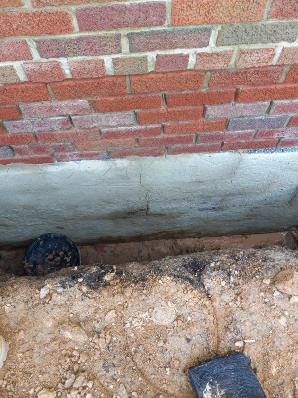 A brick building foundation with a black pipe partially buried in brown soil.