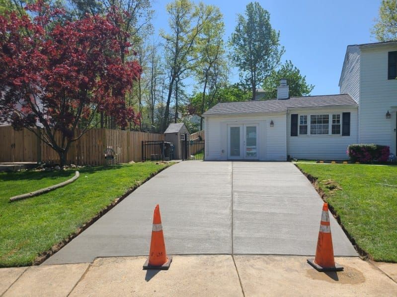 Freshly poured concrete driveway with two orange cones, in front of a white house and green lawn.