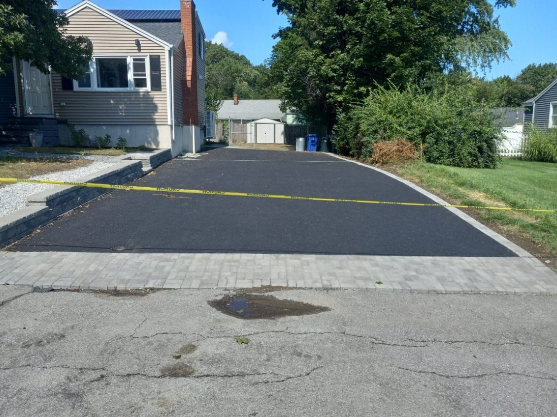 Newly paved black asphalt driveway with gray brick border in front of a beige house.