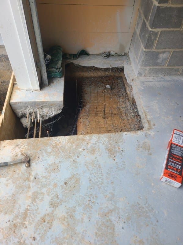 Concrete floor with a rectangular section removed, exposing rebar and soil. A support beam and cinder block wall are present.