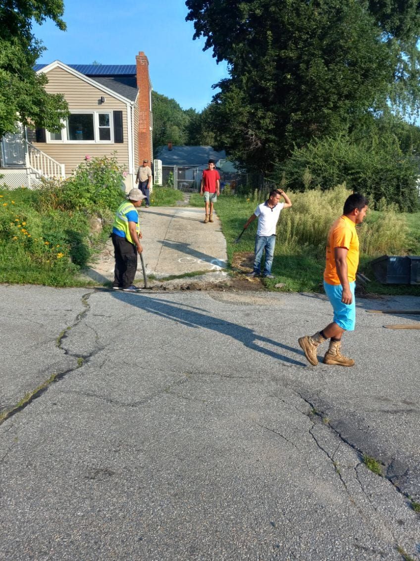 Five people working on a cracked asphalt driveway, with a house and foliage in the background.