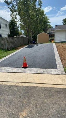 Newly paved driveway with brick border and orange cone, near a shed and houses.