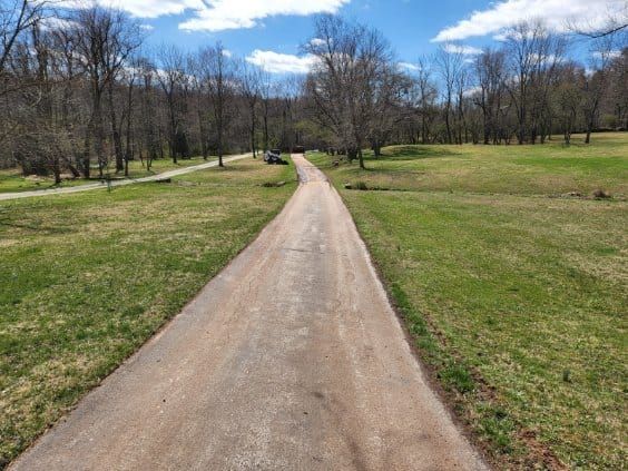 Paved road through a grassy field, leading into a wooded area under a blue sky.