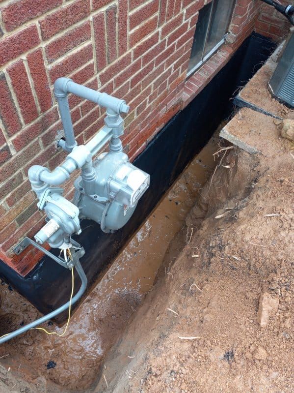 Trench dug next to a brick wall, with a gas meter installed. Black lining visible.