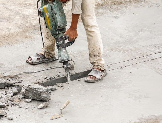 Person using a jackhammer to break concrete, outdoors. They are wearing sandals and dirty pants.