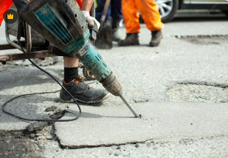 Person using a jackhammer on a cracked asphalt road. Another person in orange pants stands behind him.