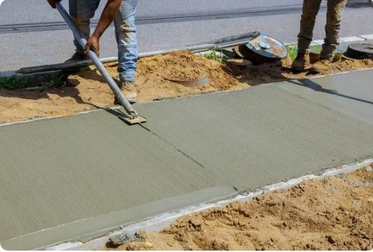 Construction worker smoothing wet concrete sidewalk with a long tool.