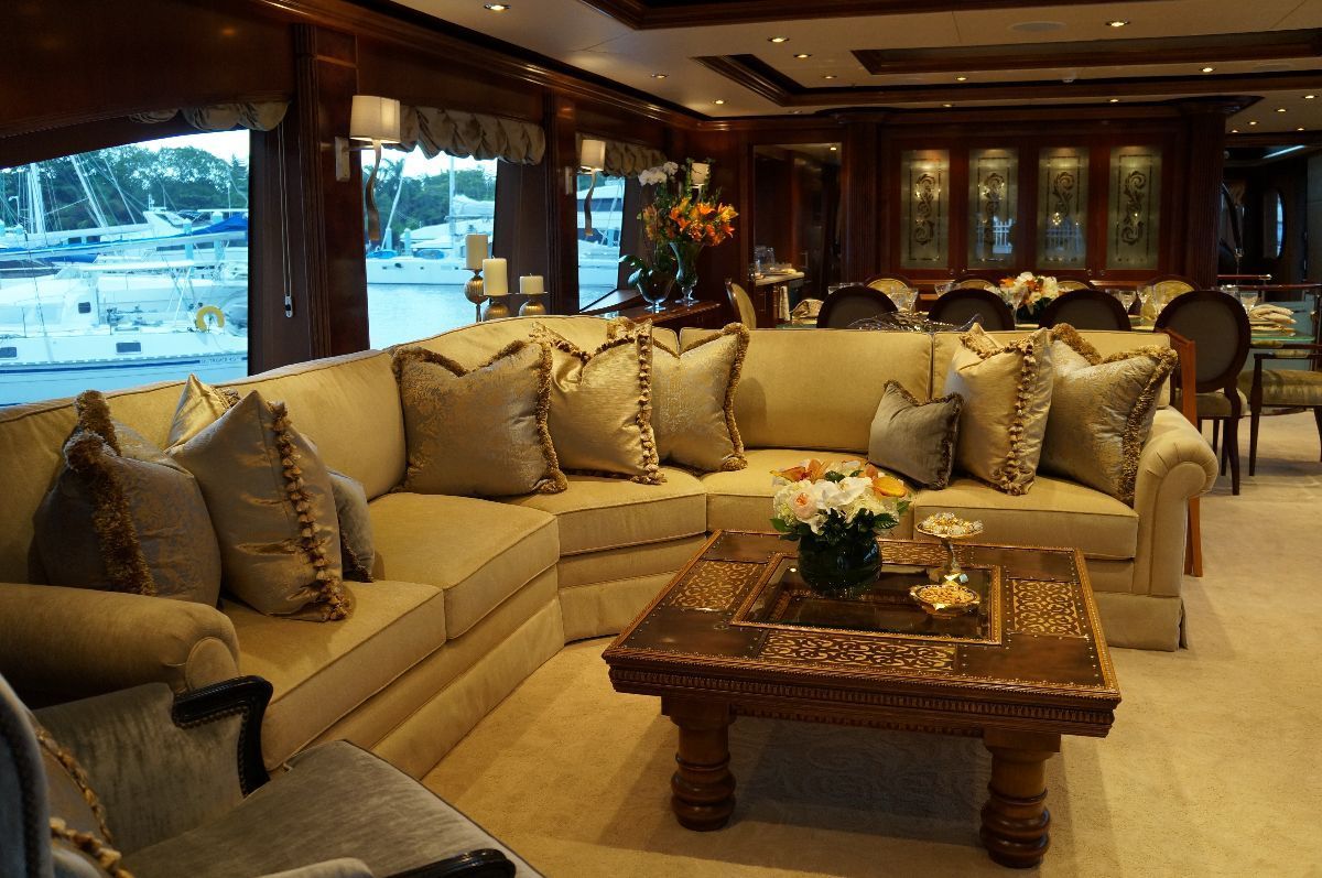 Luxurious yacht interior with beige sectional sofa, carved wooden coffee table, and large windows overlooking water.