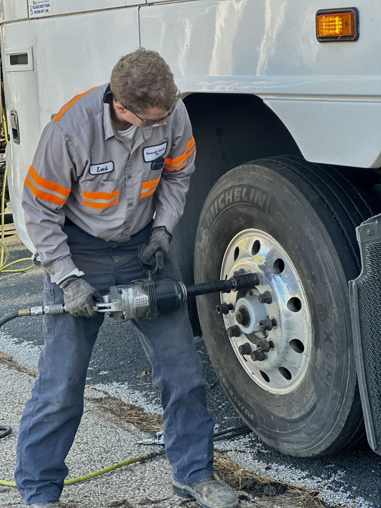 A mechanic in a work uniform using a power tool on a bus tire outdoors.