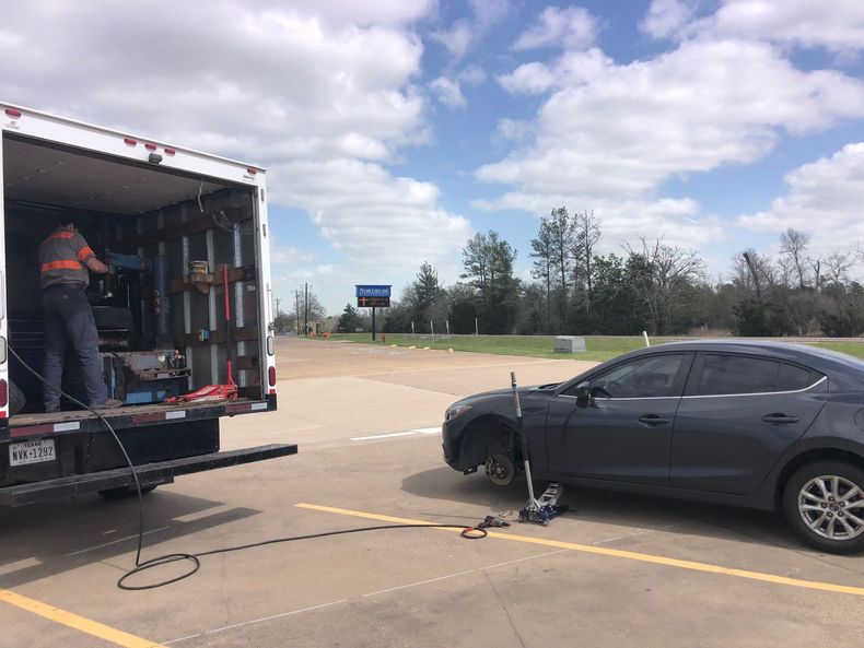 A mechanic changing a tire on a gray car in a parking lot, next to a service truck. The sky is cloudy.