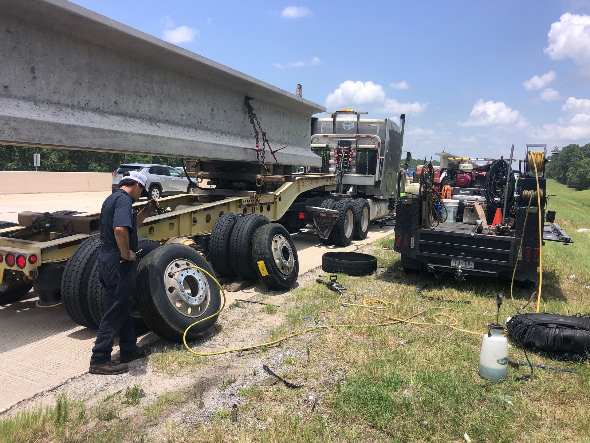 Semi-truck carrying concrete beam, roadside tire repair; workers, blue sky.