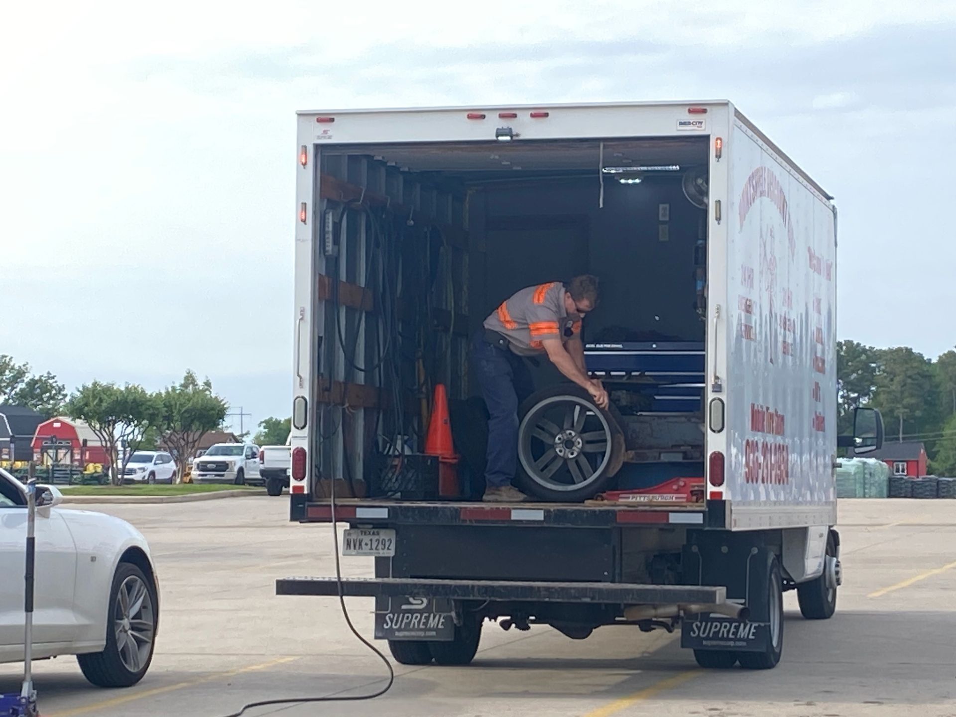 Man in truck bed changing car tire. White truck parked next to a white sports car in a parking lot.