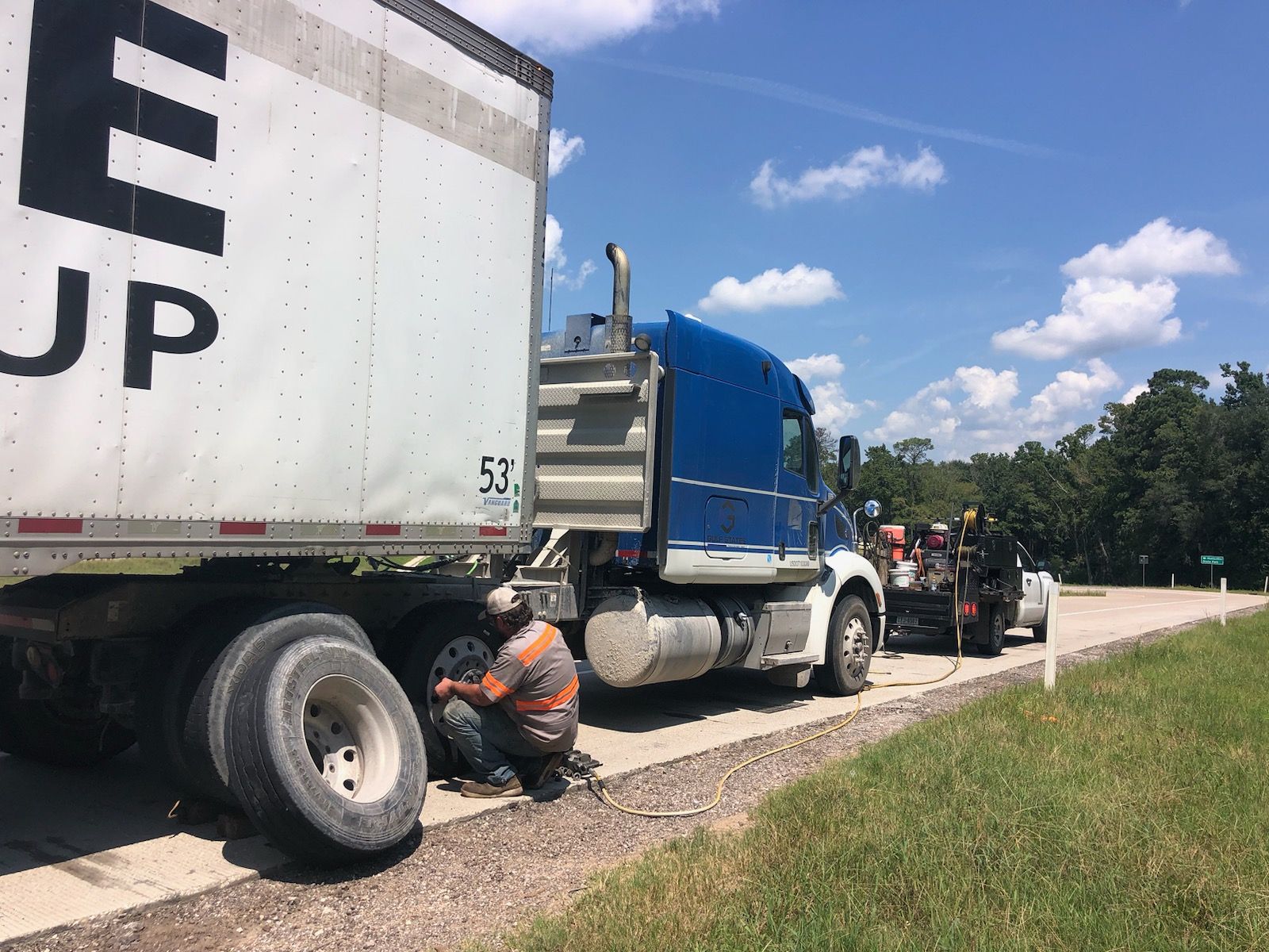 Trucker changing a tire on a semi-truck on the side of a road. Tow truck is present.