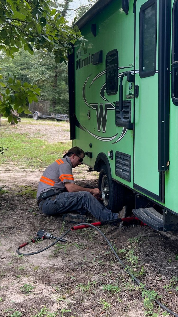 A person in work clothes changes a tire on a green camper in a grassy area.