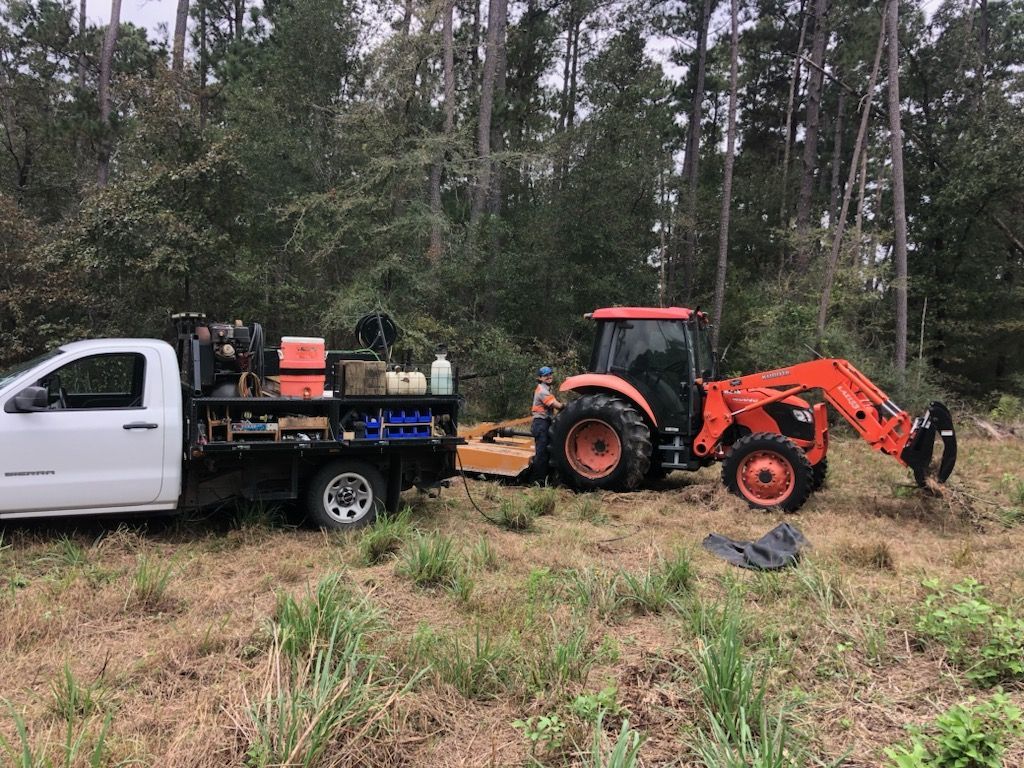 White truck with equipment and orange tractor in a grassy field near trees.